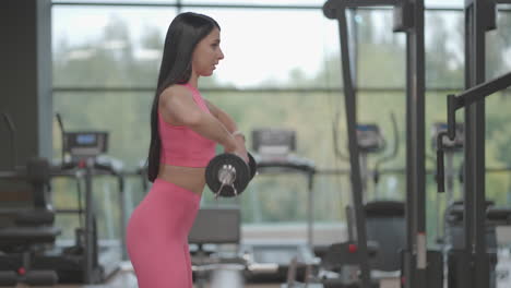 A-hispanic-brunette-woman-in-a-pink-suit-pulls-the-barbell-to-the-top-while-training-her-shoulders-in-the-gym.-Standing-exercise-for-training-the-shoulders-and-arms.-Weight-training